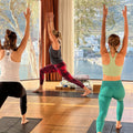 Three women practicing yoga in a room with large windows overlooking a scenic view.