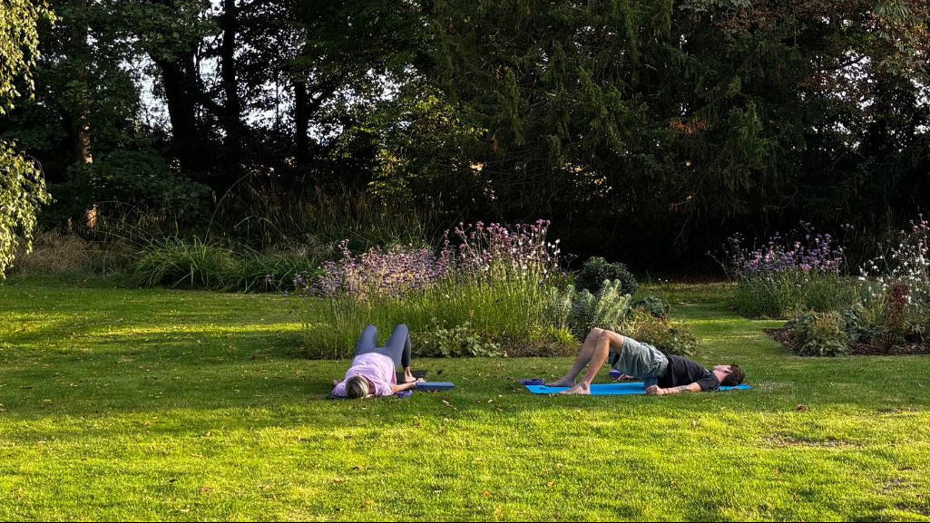Two people lying on yoga mats in a grassy area with trees and plants in the background
